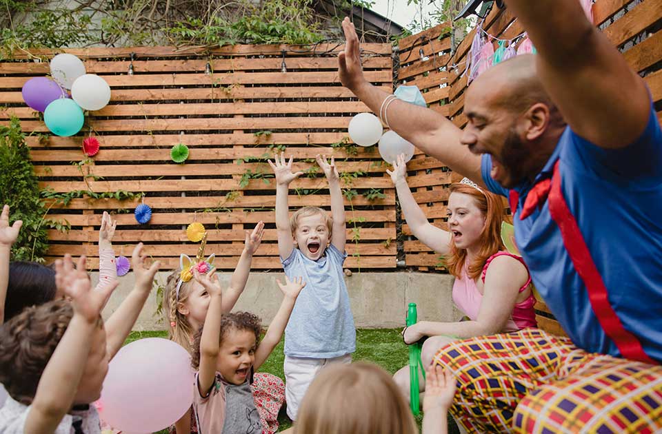 dečak sa podignutim rukama okružen decom i balonima u dvorištu