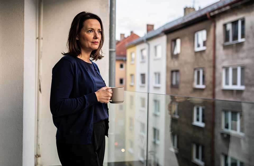 žena stoji na balkonu držeći šolju i gleda preko staklene ograde